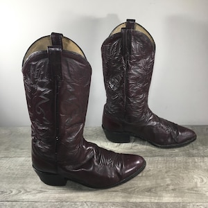 Può includere: Un paio di stivali da cowboy bordeaux in pelle. Gli stivali presentano cuciture decorative, punte a punta e tacchi impilati. Sono esposti su una superficie di legno. Gli stivali sono di un colore intenso.