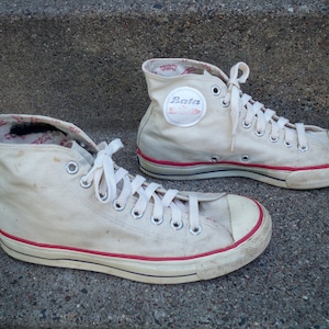 Pode incluir: Um par de t&ecirc;nis branco de lona de cano alto com detalhes vermelhos e cadar&ccedil;os brancos. Os t&ecirc;nis t&ecirc;m uma etiqueta da marca Bata no lado.