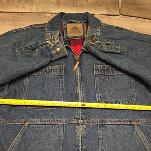 May include: A blue denim jacket with a red plaid lining, a front zipper closure, and two chest pockets. A yellow measuring tape is placed across the chest, showing a width of approximately 24 inches. The jacket features a collar and metal buttons.