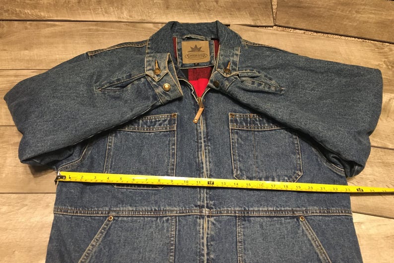 May include: A blue denim jacket with a red plaid lining, a front zipper closure, and two chest pockets. A yellow measuring tape is placed across the chest, showing a width of approximately 24 inches. The jacket features a collar and metal buttons.