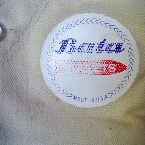 Pode incluir: Sapato branco de lona com um logotipo azul e vermelho que diz "Bata" e "Sports" com uma borda branca e o texto "Made in U.S.A."