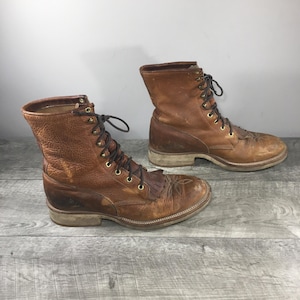 Puede incluir: Un par de botas de cuero marrón con cordones, acabado texturizado, ojales de metal dorado, cordones marrón oscuro y un detalle de flecos decorativos sobre el empeine. Las botas tienen un tacón apilado y punta redonda con costuras.