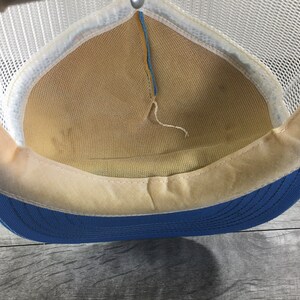 May include: The inside of a blue and white mesh trucker hat. The hat has a white mesh crown and a blue brim. The inside of the hat is lined with a beige fabric.