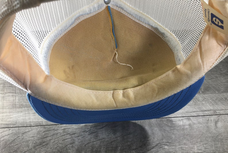 May include: The inside of a blue and white mesh trucker hat. The hat has a white mesh crown and a blue brim. The inside of the hat is lined with a beige fabric.
