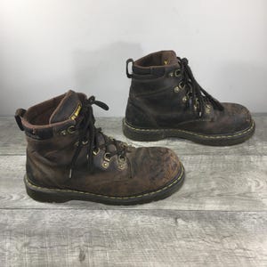 May include: A pair of dark brown leather Dr. Martens work boots featuring a high-top silhouette, black laces, metal eyelets, and a yellow stitched welt. The boots have a padded collar, a pull tab at the heel, and a rugged, worn-in appearance.