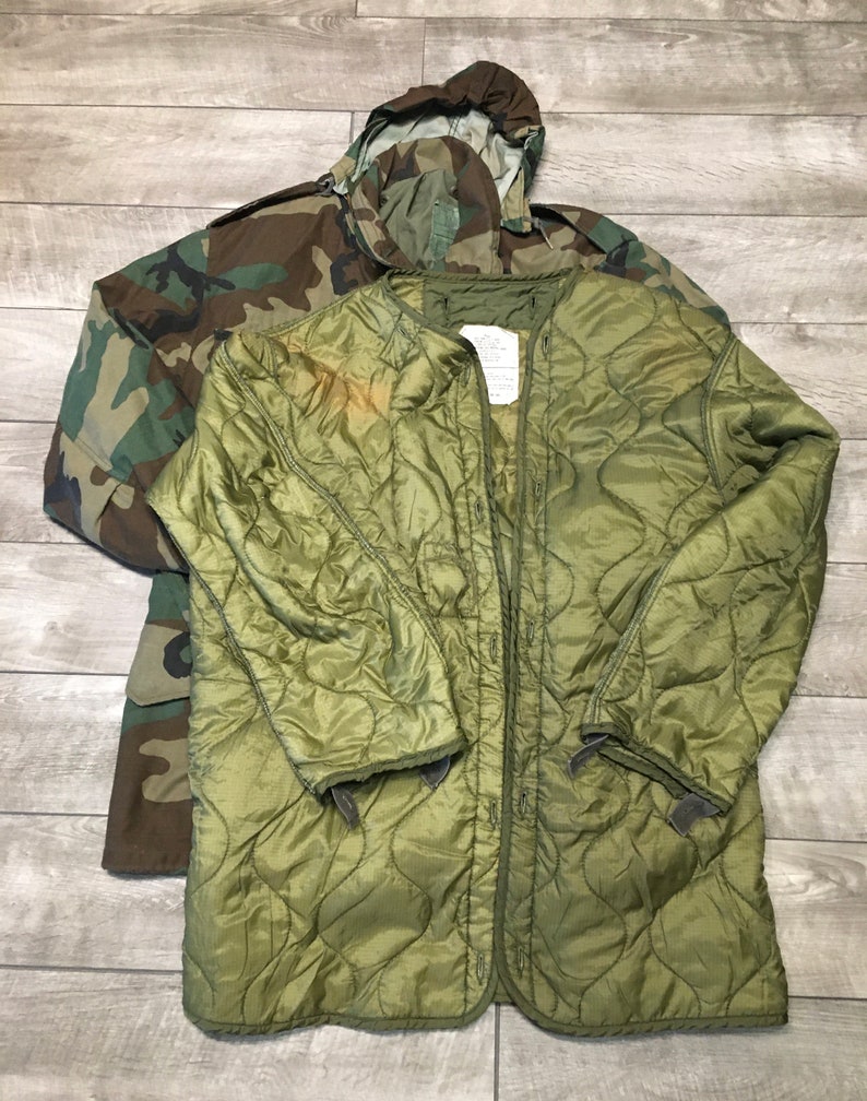 K&ouml;nnte beinhalten: Eine gr&uuml;ne gesteppte Jacke mit Druckknopfverschluss und Kapuze. Die Jacke liegt auf einem Holzboden mit einer Tarnjacke darunter.