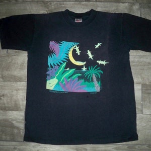 Puede incluir: Camiseta negra con un estampado gráfico colorido de una luna creciente, palmeras y lagartos. El texto "Somewhere Out There In The Ocean Blue" está impreso debajo del gráfico.