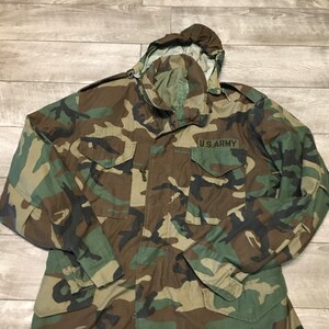 K&ouml;nnte beinhalten: Eine gr&uuml;ne, braune und schwarze Tarnjacke mit Kapuze und einem Aufn&auml;her mit der Aufschrift "U.S. ARMY".