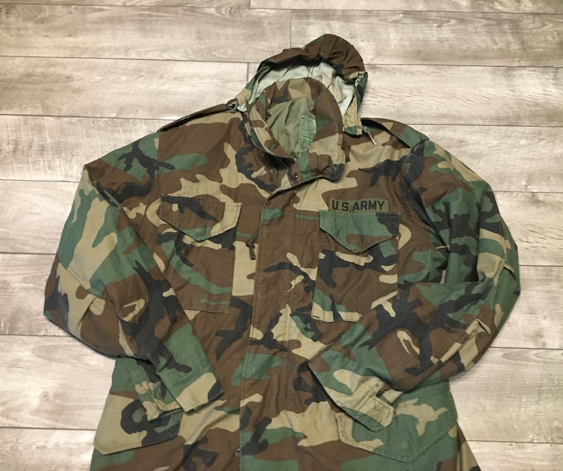 K&ouml;nnte beinhalten: Eine gr&uuml;ne, braune und schwarze Tarnjacke mit Kapuze und einem Aufn&auml;her mit der Aufschrift "U.S. ARMY".