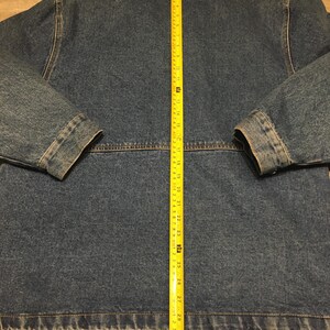 May include: A blue denim jacket with a red collar lining, featuring a yellow measuring tape placed vertically down the center back, showing a total length of approximately 31 inches from the collar seam to the bottom hem of the garment.
