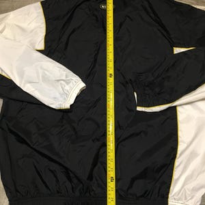 May include: Black and white nylon jacket with yellow trim. The jacket has a zipper closure and a white collar. The sleeves are white, and the body is black. The jacket is measured with a yellow tape measure.