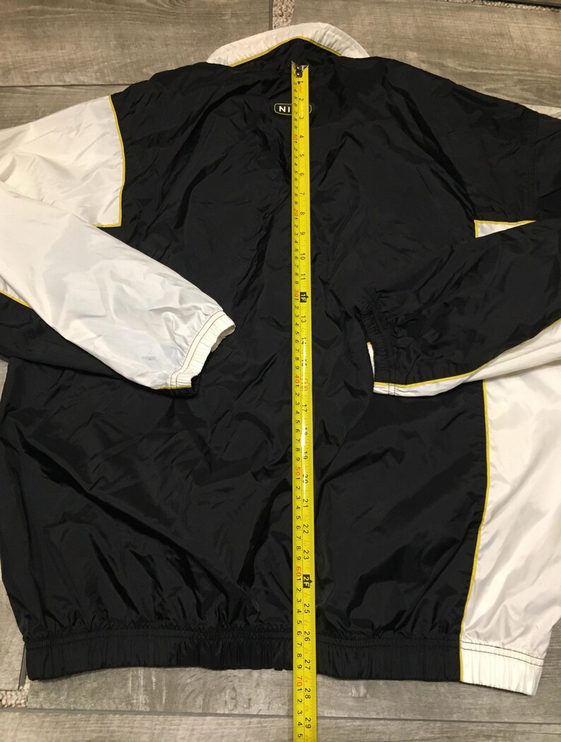 May include: Black and white nylon jacket with yellow trim. The jacket has a zipper closure and a white collar. The sleeves are white, and the body is black. The jacket is measured with a yellow tape measure.