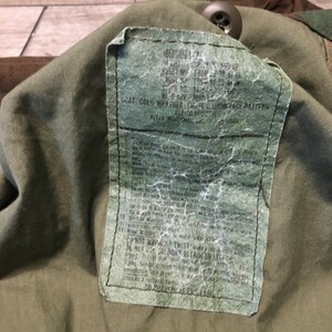 K&ouml;nnte beinhalten: Nahaufnahme eines gr&uuml;nen Milit&auml;rjacken-Etiketts mit Pflege- und Gebrauchsanweisungen. Das Etikett enth&auml;lt den Text "GOAT, COLD WEATHER, CHATE CAMOUFLAGE PATTERN" und "ALPHA INDUSTRIES, INC."