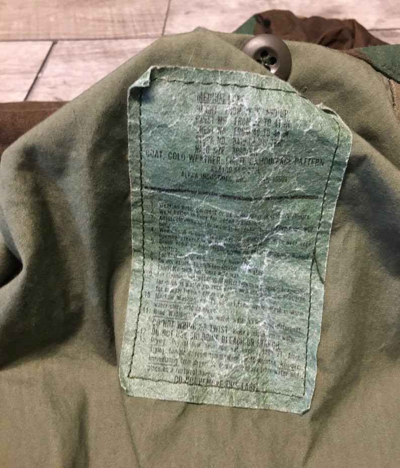 K&ouml;nnte beinhalten: Nahaufnahme eines gr&uuml;nen Milit&auml;rjacken-Etiketts mit Pflege- und Gebrauchsanweisungen. Das Etikett enth&auml;lt den Text "GOAT, COLD WEATHER, CHATE CAMOUFLAGE PATTERN" und "ALPHA INDUSTRIES, INC."
