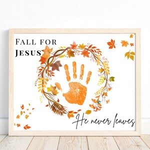 Puede incluir: Impresión enmarcada con las palabras "FALL FOR JESUS" y "He never leaves". La obra presenta una huella de mano naranja rodeada por una corona de hojas de otoño. El marco es de color claro.