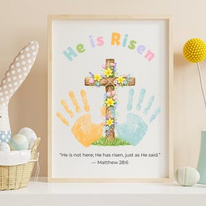 May include: A framed print with the words "He is Risen" in pastel colours. A wooden cross is decorated with flowers, and there are handprints in orange and blue. The quote "He is not here; He has risen, just as He said." is below.