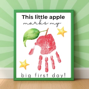 May include: A framed print with a green border features a red handprint shaped like an apple, with a green leaf and stem. The text reads "This little apple marks my big first day!" with two yellow star accents.