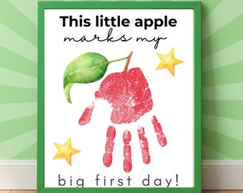 May include: A framed print with a green border features a red handprint shaped like an apple, with a green leaf and stem. The text reads "This little apple marks my big first day!" with two yellow star accents.
