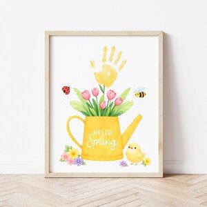 Puede incluir: Una impresi&oacute;n en acuarela enmarcada con una regadera amarilla llena de tulipanes rosas y hojas verdes. Una huella de mano amarilla est&aacute; encima de las flores. Tambi&eacute;n se incluyen una mariquita, una abeja y un pollito. El texto "Hello Spring".