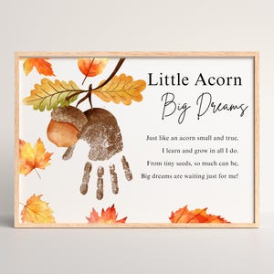 Puede incluir: Impresión enmarcada con el texto "Little Acorn Big Dreams" y un poema. La obra presenta una bellota, hojas de otoño y una huella de mano. El marco es de madera clara.