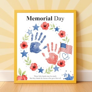 May include: A framed print with a yellow border. The artwork features a wreath of red poppies, blue stars, and green leaves around two handprints, one blue and one red. The American flag and text "Memorial Day" are also included.
