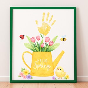 Puede incluir: Una impresi&oacute;n enmarcada con un borde verde. La obra de arte presenta una regadera amarilla llena de tulipanes rosas y hojas verdes. Una huella de mano amarilla est&aacute; encima de la regadera, con una mariquita y una abeja. Las palabras "Hello Spring" est&aacute;n escritas en la regadera.