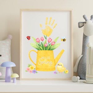 Puede incluir: Una impresi&oacute;n enmarcada con una ilustraci&oacute;n en acuarela de una regadera amarilla llena de tulipanes rosas y hojas verdes. Una huella de mano amarilla est&aacute; encima de la regadera. Las palabras "Hello Spring" est&aacute;n en la regadera. Una mariquita, una abeja y un pollito tambi&eacute;n est&aacute;n en la imagen.