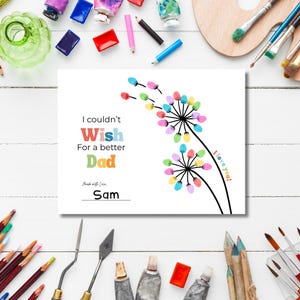 May include: A white greeting card with the text "I couldn't Wish For a better Dad" and the name "Sam". The card features a colorful dandelion design with the words "I love you!" on the side. Paint tubes and brushes surround the card.