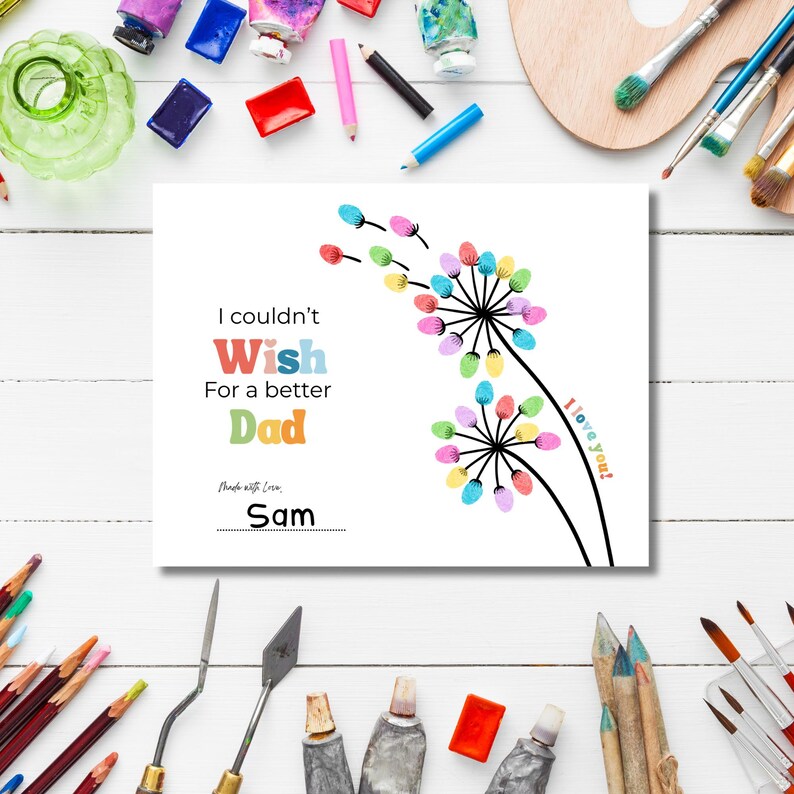 May include: A white greeting card with the text "I couldn't Wish For a better Dad" and the name "Sam". The card features a colorful dandelion design with the words "I love you!" on the side. Paint tubes and brushes surround the card.