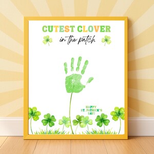 Könnte beinhalten: Ein gerahmter Druck mit gelbem Rahmen. Er zeigt einen grünen Handabdruck und den Text "Cutest Clover in the Patch". Aquarell-Kleeblätter und der Text "Happy St. Patrick's Day!" sind ebenfalls enthalten. Der Druck befindet sich auf weißem Hintergrund.