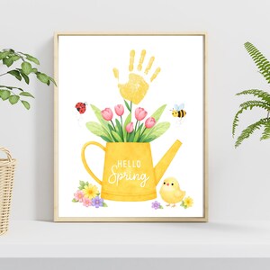 Puede incluir: Una impresi&oacute;n enmarcada con acuarela que presenta una regadera amarilla llena de tulipanes rosas y hojas verdes. Una huella de mano amarilla est&aacute; encima de la regadera. Las palabras "Hello Spring" est&aacute;n escritas en la regadera. Tambi&eacute;n se muestra una mariquita, una abeja y un pollito.