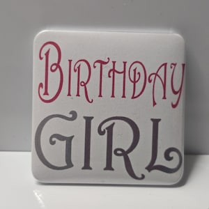 May include: Square white button with the words "BIRTHDAY GIRL" in a decorative font. The word "BIRTHDAY" is in a dark pink color, and the word "GIRL" is in a dark gray color. The button is on a white surface.