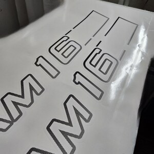 May include: Two black and grey M16 decals are visible against a white backdrop. The decals feature a textured design and are partially covered by a transparent protective layer. The M16 text is in a bold, outlined font.