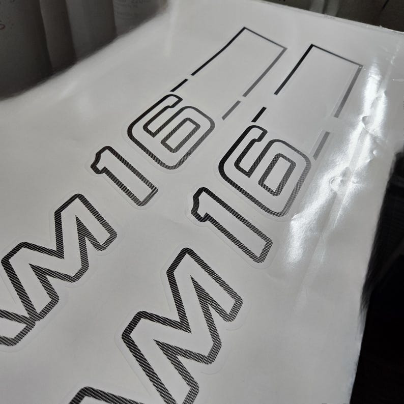 May include: Two black and grey M16 decals are visible against a white backdrop. The decals feature a textured design and are partially covered by a transparent protective layer. The M16 text is in a bold, outlined font.