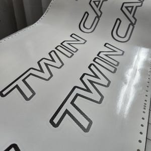 May include: A close-up shot of a sheet of white stickers. The stickers feature the words "TWIN CAM" in a bold, black font with a cross-hatch pattern. The sheet has perforated edges.
