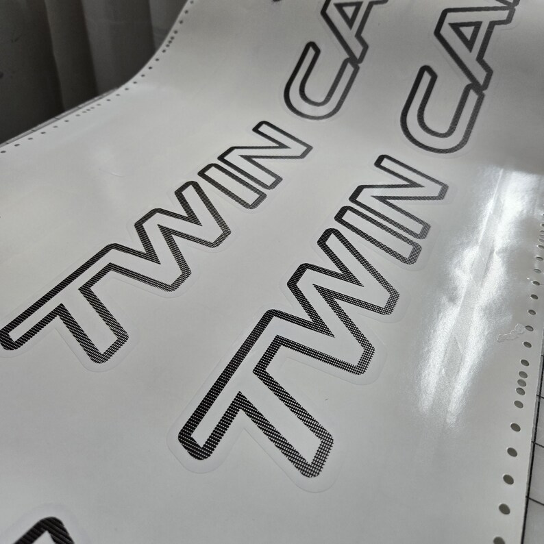 May include: A close-up shot of a sheet of white stickers. The stickers feature the words "TWIN CAM" in a bold, black font with a cross-hatch pattern. The sheet has perforated edges.
