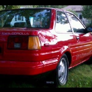 May include: A red Toyota Corolla coupe parked on grass. The car has a shiny finish, silver wheels, and a black side mirror. The text "TOYOTA COROLLA" is visible on the rear.