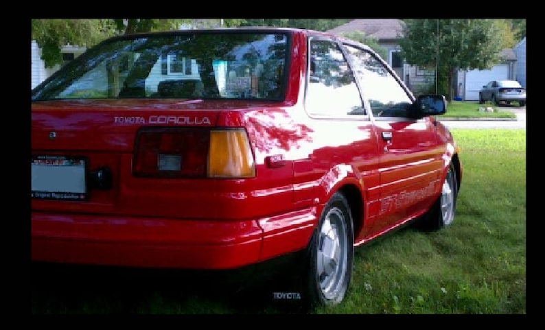 May include: A red Toyota Corolla coupe parked on grass. The car has a shiny finish, silver wheels, and a black side mirror. The text "TOYOTA COROLLA" is visible on the rear.