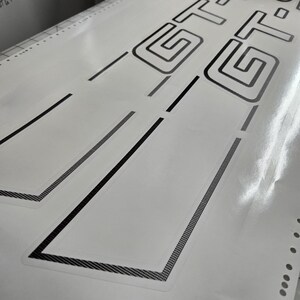 May include: White sheet featuring black GT logo and accent lines. The GT logo is a geometric design. The accent lines have a textured, dotted pattern. The sheet has a glossy finish, likely a decal or sticker for a vehicle.