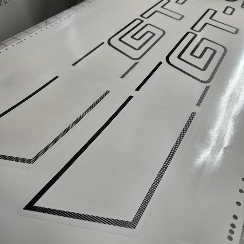 May include: White sheet featuring black GT logo and accent lines. The GT logo is a geometric design. The accent lines have a textured, dotted pattern. The sheet has a glossy finish, likely a decal or sticker for a vehicle.