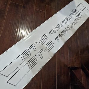 May include: White decal featuring the text "GT-S TWIN CAM 16" in a black, outlined font. The decal has a rectangular shape with angled lines on the left. It is on a dark wooden surface.