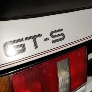 May include: Close-up of a silver Toyota GT-S car. The GT-S logo is prominently displayed on the trunk. The car has red tail lights and a California license plate. The Toyota logo is visible.