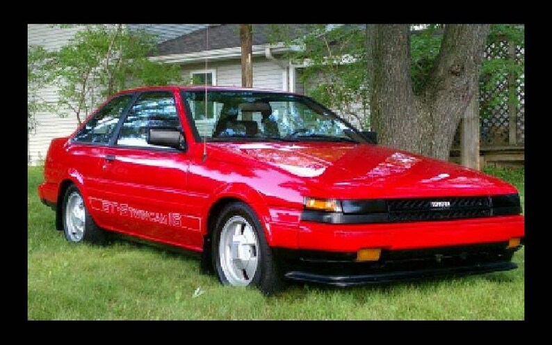 May include: A vibrant red Toyota sports car with black detailing, parked on grass. The car has a sleek design, visible headlights, and the text "GT-S Sprinter" on the side. A classic car.