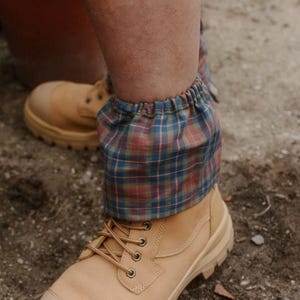 May include: A pair of tan leather work boots featuring a lace-up front and a thick rubber sole, worn with a blue, red, and green plaid fabric gaiter or cuff accessory that wraps around the ankle area with an elasticised top edge.