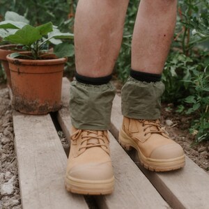 May include: A pair of tan work boots with green gaiters. The boots are being worn by a person standing on a wooden walkway. The person is wearing the boots in a garden setting.