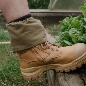 May include: A pair of tan work boots with a zip on the side and a brown gaiter. The boots are being worn by someone standing on a wooden surface.