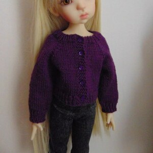 Puede incluir: Una muñeca con un cárdigan de punto morado con botones y pantalones grises.