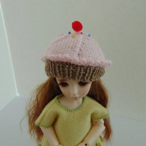 May include: A doll wearing a pink and brown knitted cupcake hat. The hat has a red cherry on top and colorful sprinkles.