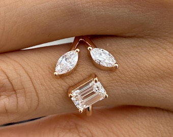 May include: A rose gold open ring featuring two marquise-cut clear stones arranged in a V-shape at the top and one rectangular emerald-cut clear stone positioned below, creating a bypass design that rests on the finger.
