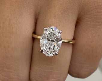 May include: A gold band engagement ring featuring a large, oval-cut diamond. The diamond is set with four prongs, and the band has a simple, polished design. The ring is shown on a finger.